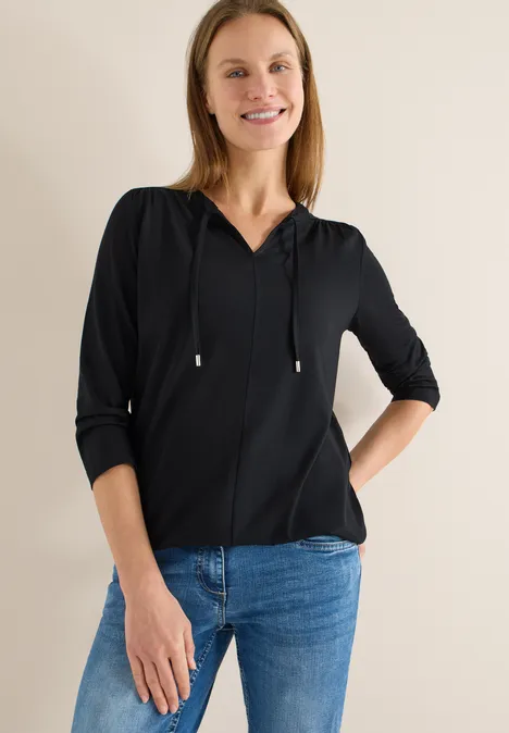 Frau in schwarzer Kapuzenbluse und blauer Jeans steht mit einer Hand in der Tasche.